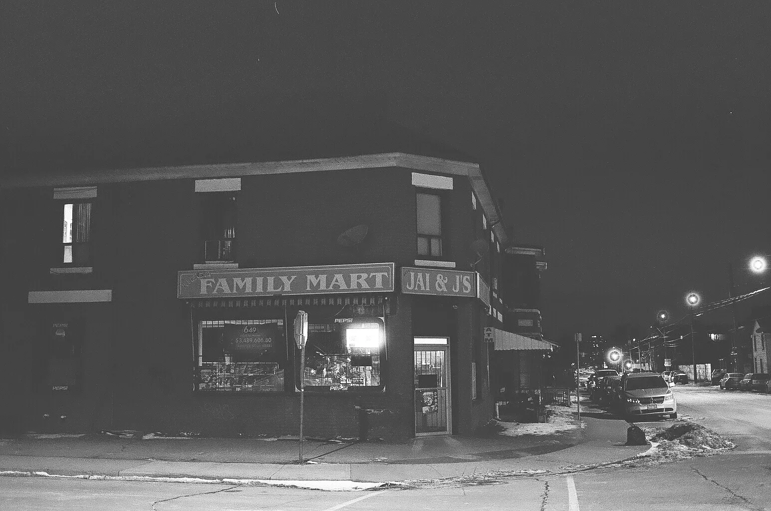 A black and white photo of a corner store at night in the snow.